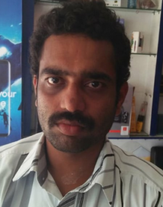 Photo of a man wearing a white striped shirt. He has brown skin and dark-brown eyes; his hair is dark-brown, and he has a mustache and some beard stubble. His expression is neutral.