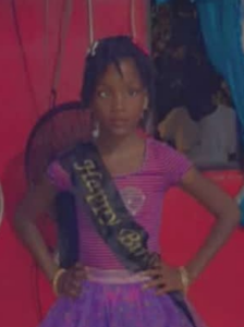 Photo of a small girl with a mop of curly black hair and brown skin; she is wearing a pink T-shirt with a black sash. She has her hands on her hips and is looking at the camera.