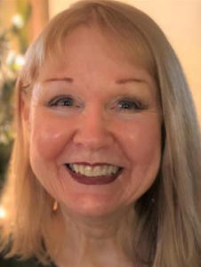 Photo of a blond woman with light skin; she is smiling widely. Her lips are done in dark-red lipstick.