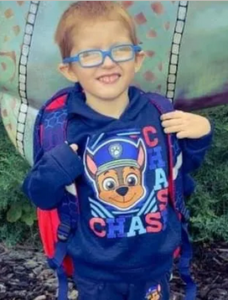 Photo of a little boy with short red hair and pale skin, wearing a backpack, a blue Paw Patrol shirt, and blue glasses. He is smiling for the camera, eyes squinting closed against the light.