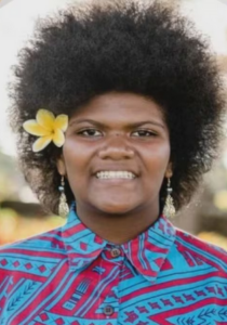 Photo of a girl with a fluffy afro and brown skin. She has a yellow flower tucked behind her ear, and is wearing a turquoise and pink blouse. She is smiling for the camera.