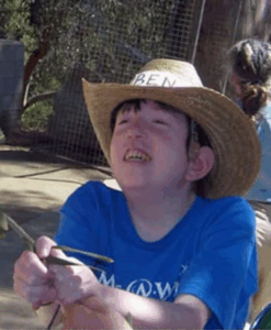 Photo of a fair-skinned, brown-haired teenage boy wearing a straw cowboy hat with a paper hatband that says, "Ben". He is smiling, looking up and to the left and holding something out; his T-shirt is blue.