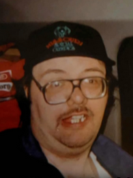 Photo of a middle-aged man in glasses and a black baseball cap printed with a logo. He has light skin and a scruffy, short mustache.