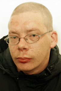 Photo of a fair-skinned young man with his hair shaved very close to the head; he is wearing small oval-rimmed glasses and a black puffy coat. His expression is neutral.