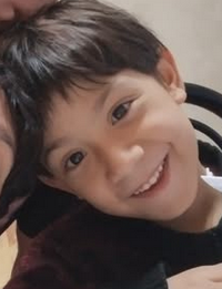 Photo of a young boy with dark-brown hair and eyes and light skin. He is smiling. He is leaning his head against the heads of two other people off the frame of the picture.