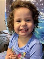 Photo of a small girl with curly brown hair and light-brown skin, wearing a blue onesie.