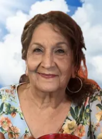 Photo of an elder woman with shoulder length brown hair and light-brown skin, smiling; she is wearing hoop earrings and a flowered blouse.