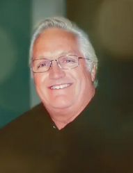 Photo of an older man with tanned skin, wearing glasses and a black shirt; his short hair is white.