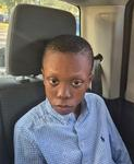 Photo of a thin preteen boy sitting in a car. He has brown skin and his black hair is cut very short; he is wearing a blue dress shirt.