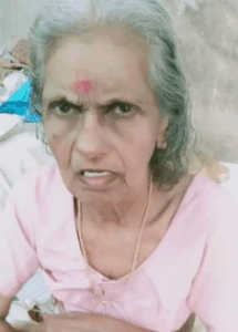 Photo of an elderly woman with shoulder-length gray hair. She has light-brown skin and a splotch of red dye as a bindi between her eyes. She is wearing a pink shirt. Her expression is confused and a little annoyed, as though the photo was taken without warning.