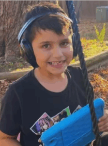 Photo of a boy with light-brown skin and brown hair, smiling for the camera. He is wearing a black T-shirt and a big pair of headphones, and holding a blue plastic communication device.