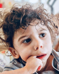 Photo of a toddler with curly, messy brown hair, deep brown eyes, and light-brown skin. He is gazing off to the left, and has stuck one index finger into his mouth.