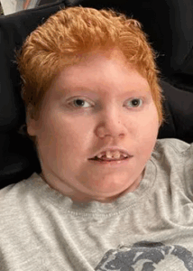 Photo of a teenage boy with light skin and short red hair, leaning against a headrest; he is wearing a gray T-shirt and has a relaxed expression on his face.