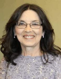 Photo of a middle-aged woman with pale skin and dark brown hair, smiling for the camera. She is wearing brown-rimmed glasses, earrings, and a gray sweater.
