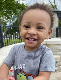 Photo of a small boy with light-brown skin and frizzy, short brown hair, smiling. He is wearing a gray T-shirt printed with a glasses-wearing dinosaur.