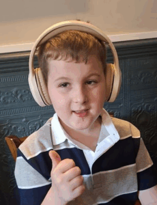 Photo of a boy with fair skin and short dark-blond hair; he is wearing a big pair of headphones and a striped polo shirt. He is smiling and giving the camera a thumbs-up.
