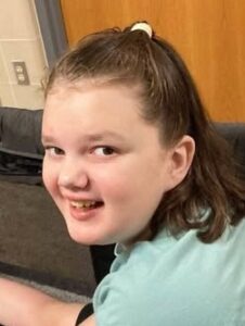 Photo of a girl with fair skin, brown hair pulled back in a half-ponytail. She is smiling, looking over her shoulder at the camera. She is wearing a mint-green shirt.