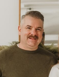 Photo of a middle-aged man, fair-skinned with a mustache and short, graying brown hair; he is smiling for the camera.