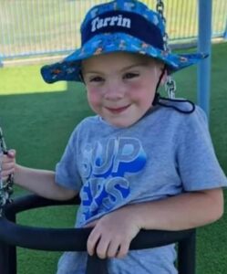 Photo of a small boy sitting in a swing. He has pale skin, and is wearing a blue sun hat with the name "Tarrin" on the band, and a gray T-shirt printed with "Sup, Peeps". He is smiling.