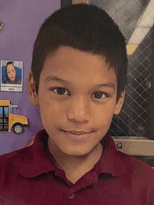 Photo of a brown-haired, brown-eyed boy with tan skin, wearing a red polo shirt. He is smiling. A school door is in the background.