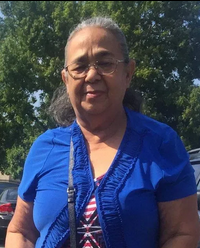 Photo of a middle-aged Hispanic woman with tan skin and graying hair pulled back in a ponytail. She is wearing glasses and a blue blouse.