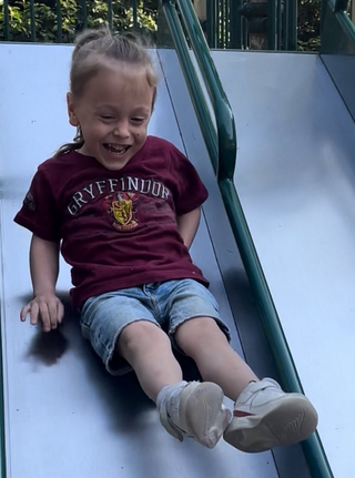Photo of a boy sliding down a slide. He has fair skin and blond hair in a ponytail; he is wearing a red shirt and denim shorts. 