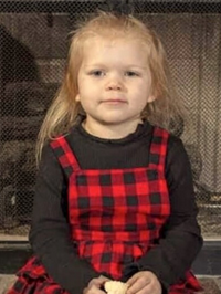 Photo of a small girl with long blond hair and pale skin. She is wearing a black sweater under a red-and-black strappy dress.